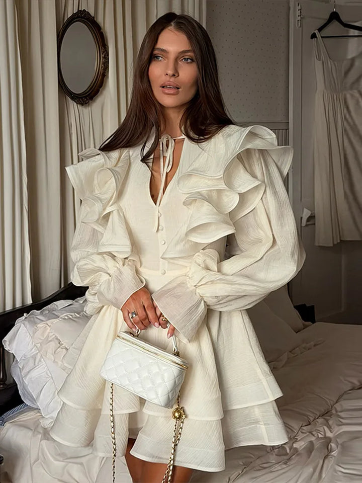 Woman in a white ruffled dress holding a white handbag in a room with curtains and a mirror.