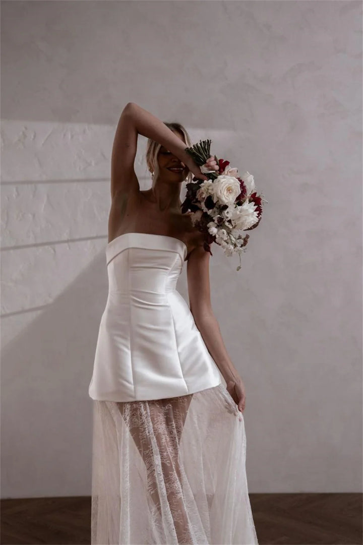 A woman modeling a strapless white wedding dress with structured side panels and a sheer tulle overskirt, holding a bouquet of white and burgundy flowers against a textured light gray wall.