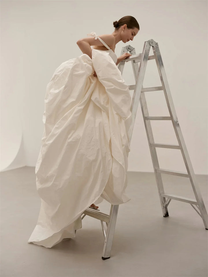 A model in a white bridal gown stands on a silver metal ladder in a minimalist studio setting, holding a small bouquet of white flowers.