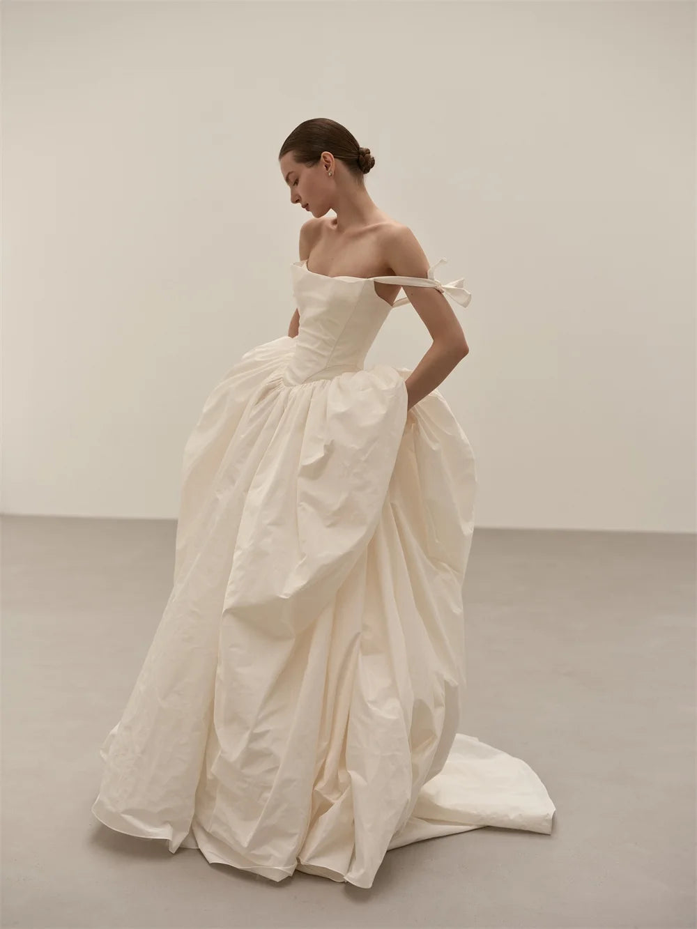 A model wearing a voluminous off-the-shoulder white wedding gown with a structured bodice and a long train, standing in a minimalist studio with a light gray floor and off-white wall.