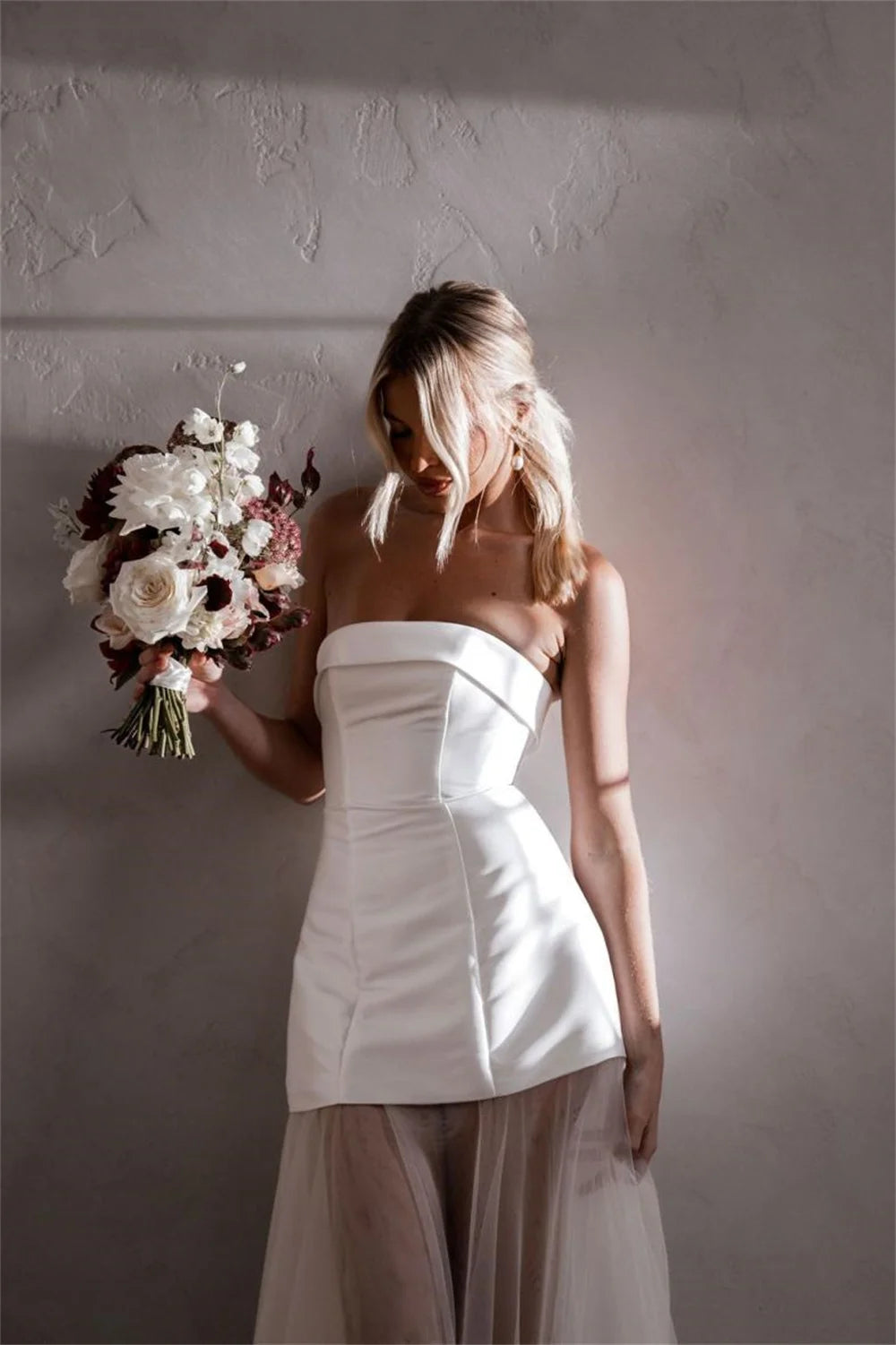 A woman wearing a strapless white corset-style dress with a sheer blush tulle skirt, holding a bouquet of white and burgundy flowers against a textured gray wall.