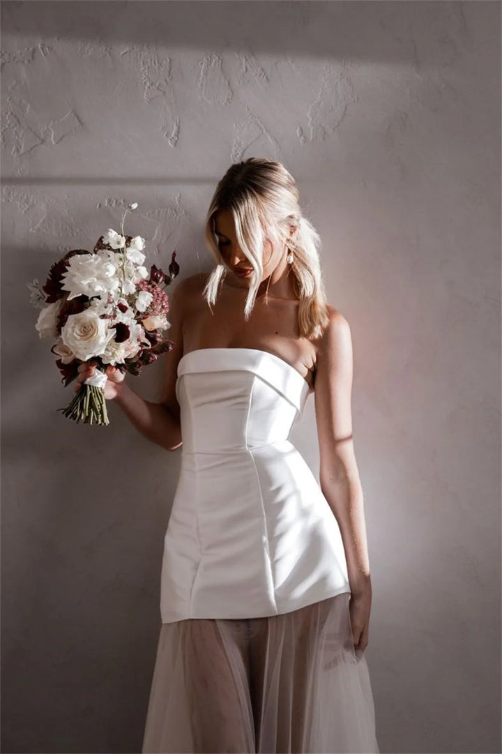 A woman wearing a strapless white corset-style dress with a sheer blush tulle skirt, holding a bouquet of white and burgundy flowers against a textured gray wall.
