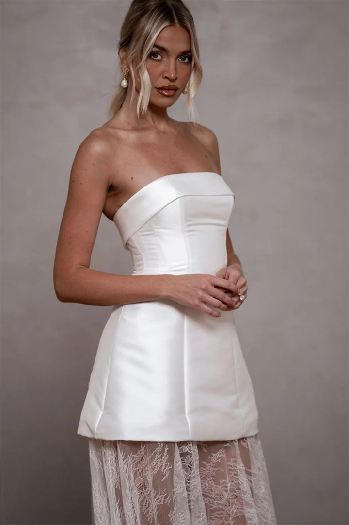 A woman modeling a strapless white satin mini dress with a structured corset-style bodice and a sheer lace overlay skirt, posed against a neutral gray studio backdrop.