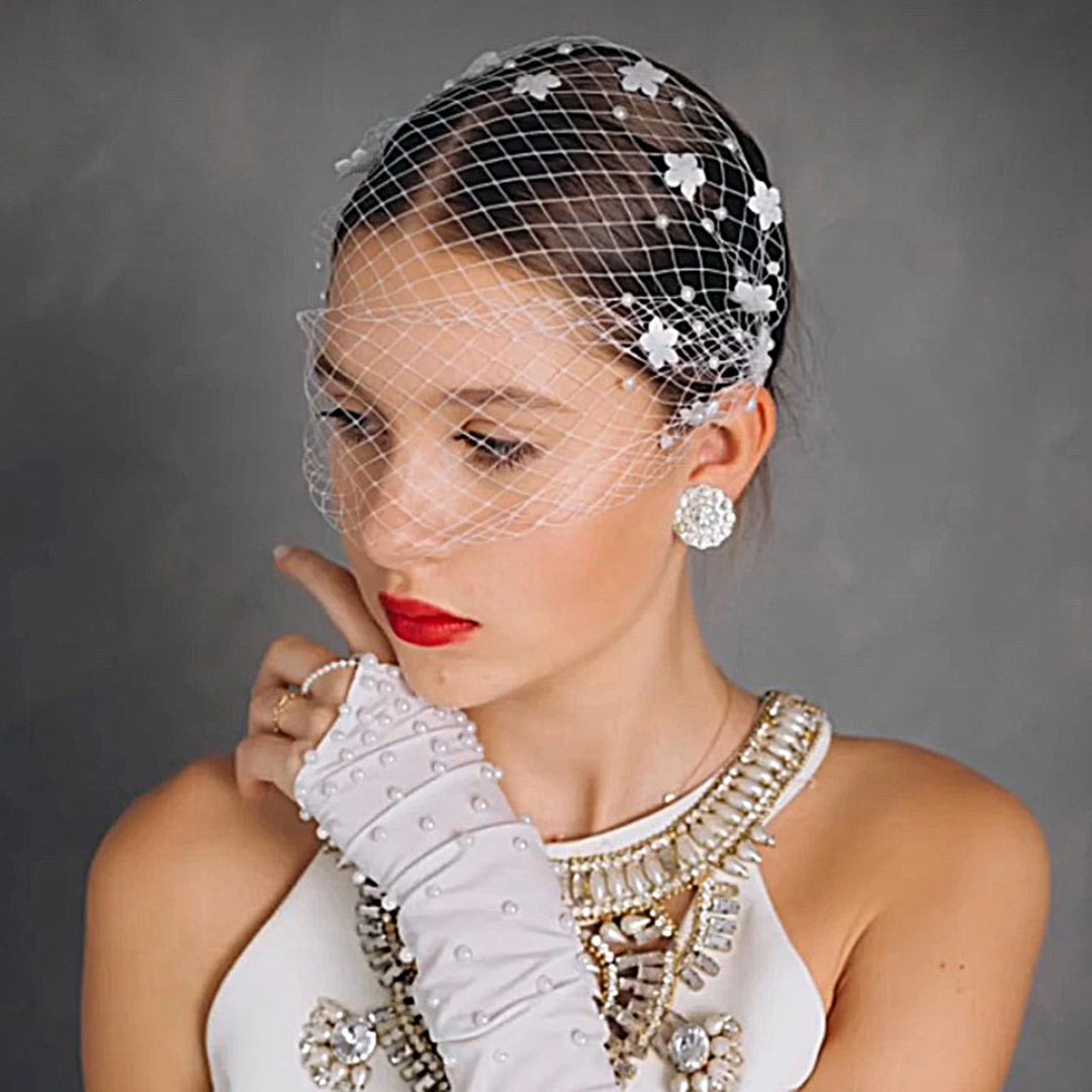 Woman wearing a decorative headpiece with flowers and a veil, against a gray background.