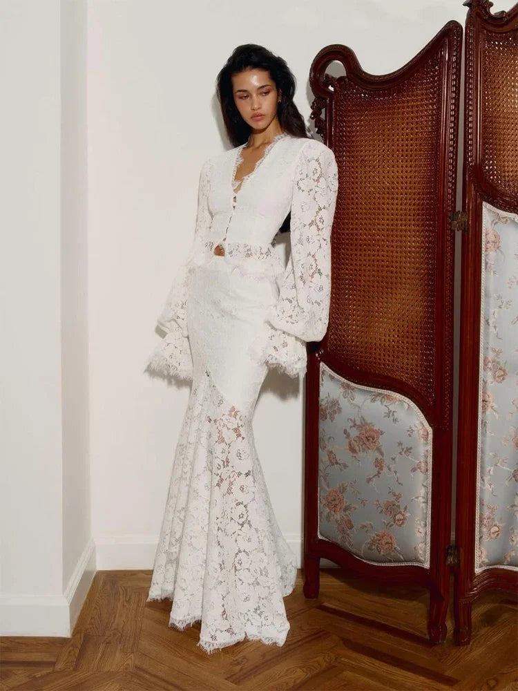 Woman in a white lace dress standing next to a decorative screen in an elegant room.