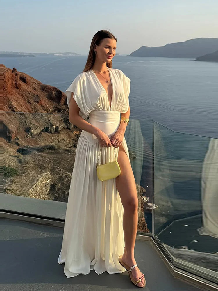 Woman in a white dress standing on a balcony with a scenic view of the ocean and mountains.