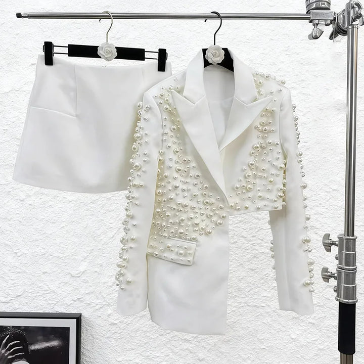 White blazer with pearl embellishments on a rack against a textured white wall.