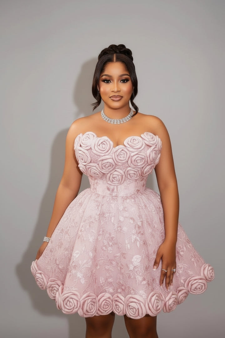 A woman modeling a light pink strapless mini dress with 3D rose applique on the bodice and flared skirt, set against a plain gray studio background.