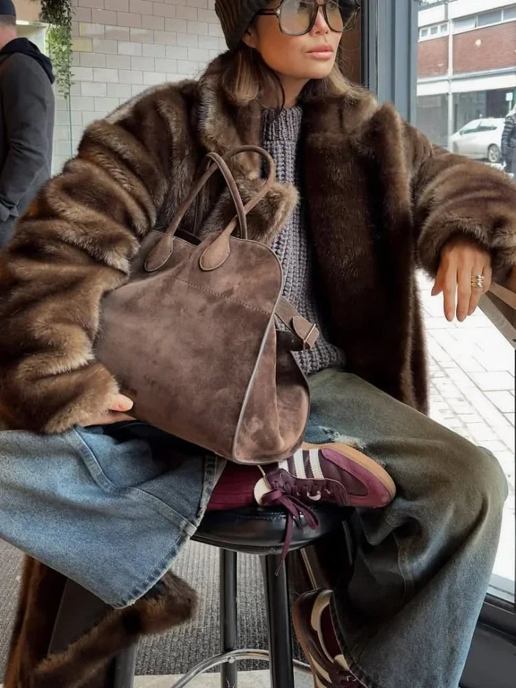 Person wearing a fur coat and holding a brown handbag, sitting on a stool.