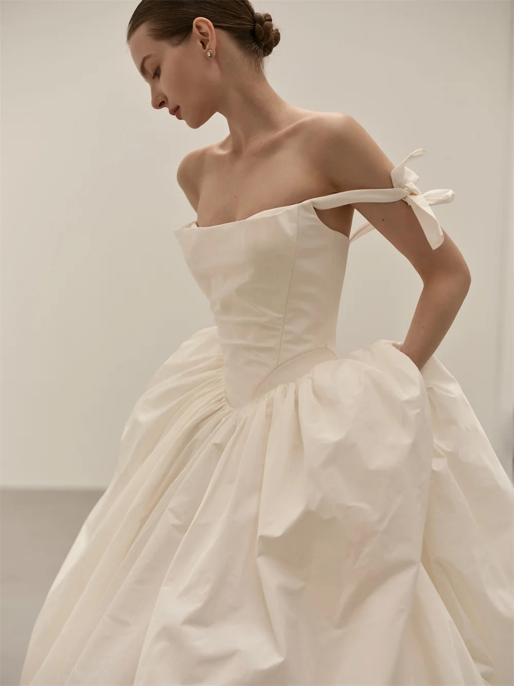 A woman wearing an off-the-shoulder white taffeta ball gown with a structured bodice and voluminous skirt, photographed in a minimalist studio setting.