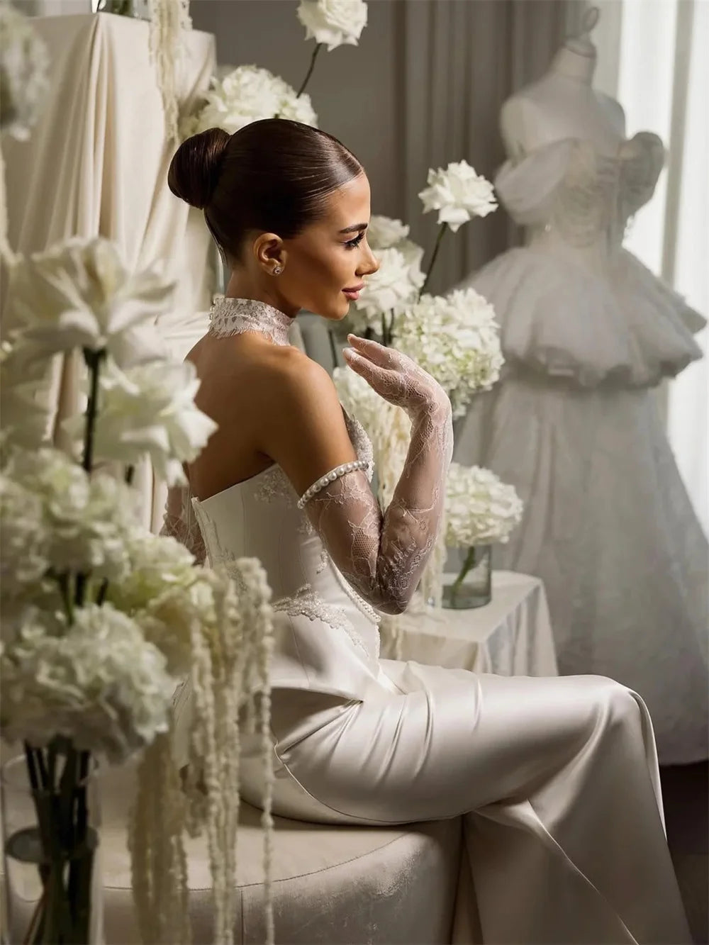 Woman in a white bridal gown sitting among flowers with wedding dresses in the background