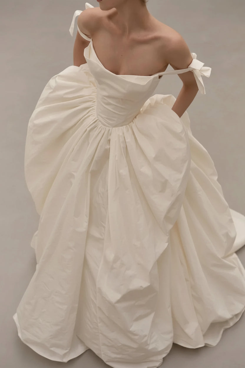 An off-the-shoulder white ballgown with a voluminous gathered skirt and bow details on the straps, shown on a model against a neutral studio background. The brand name &