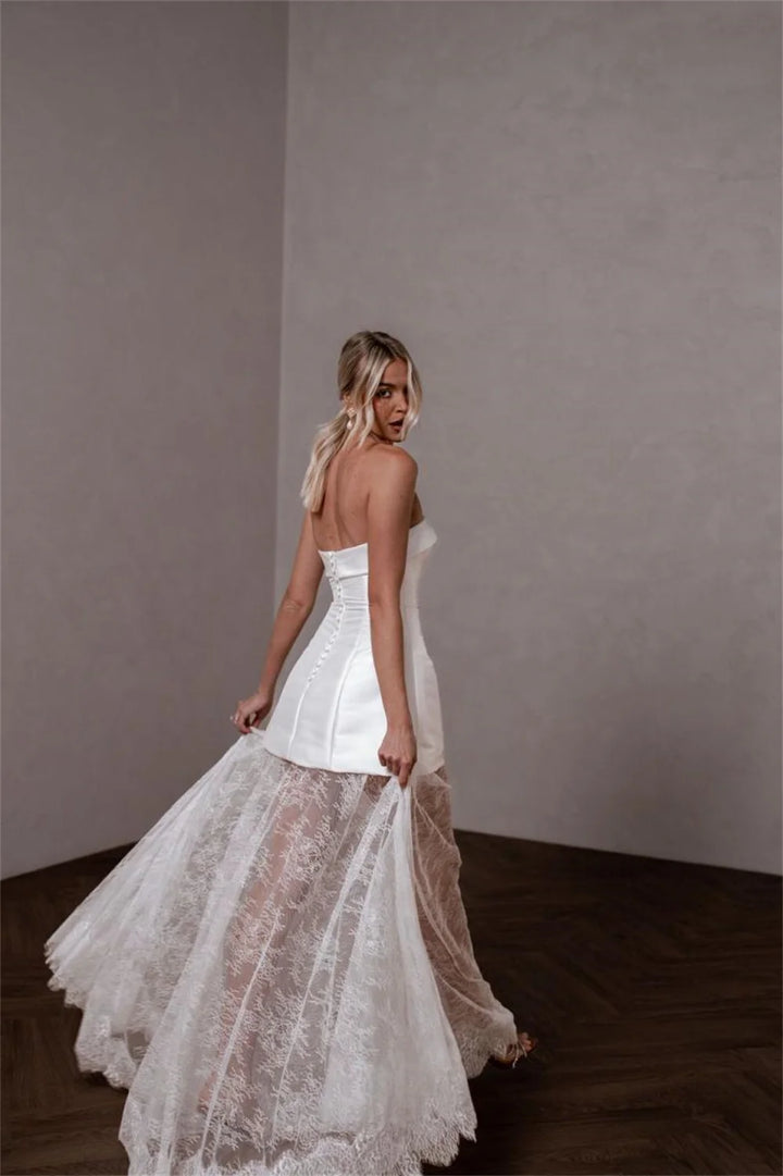 A woman wearing a strapless white wedding dress with a structured bodice and a layered tulle overskirt, photographed in a minimalist room with textured gray walls and dark wood flooring.