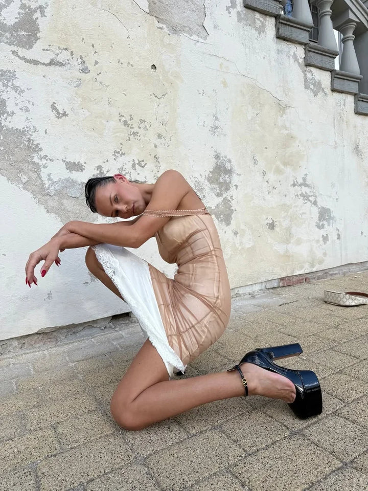 A model kneels on a stone-paved ground against a weathered plaster wall, wearing a sheer nude mesh mini dress with a white underlayer and black high-heeled platform shoes.