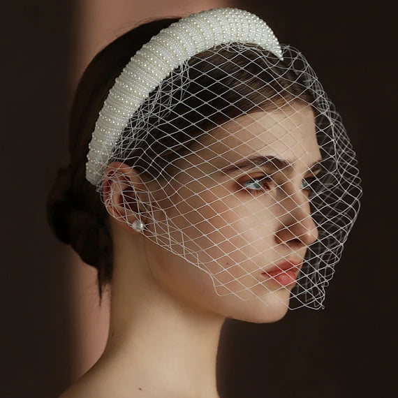 Woman wearing a white beaded headband and birdcage veil against a dark background