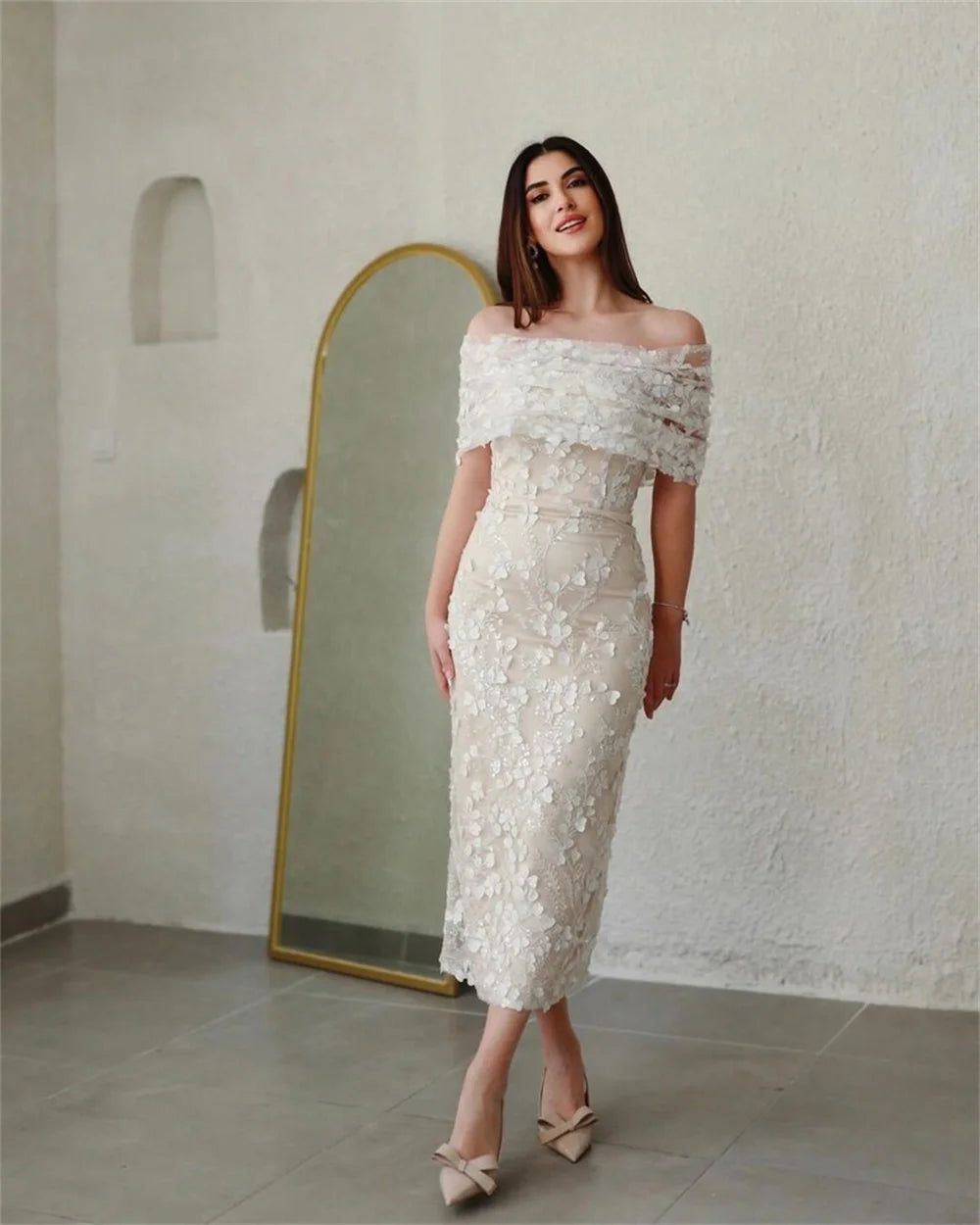 A woman standing in an elegant setting, wearing an off-white off-shoulder maxi dress with lace embroidery.
