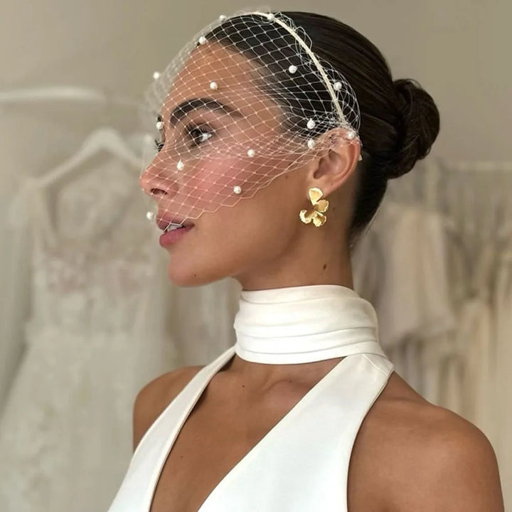 Woman wearing a white dress with a birdcage veil and gold earrings.