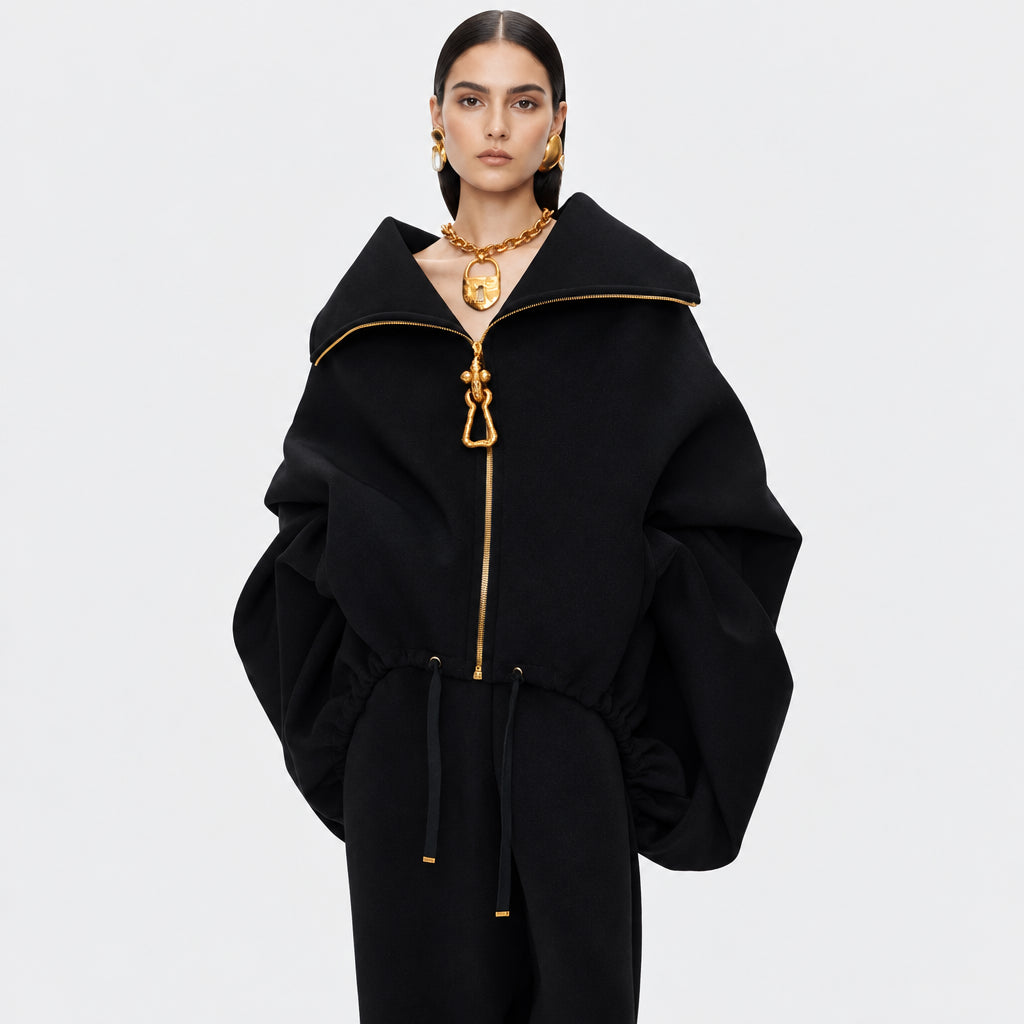 A model wearing a black oversized zip-up jacket with dramatic puffed sleeves, a high collar, and prominent gold-tone hardware including a decorative zipper pull and gold chain necklace.