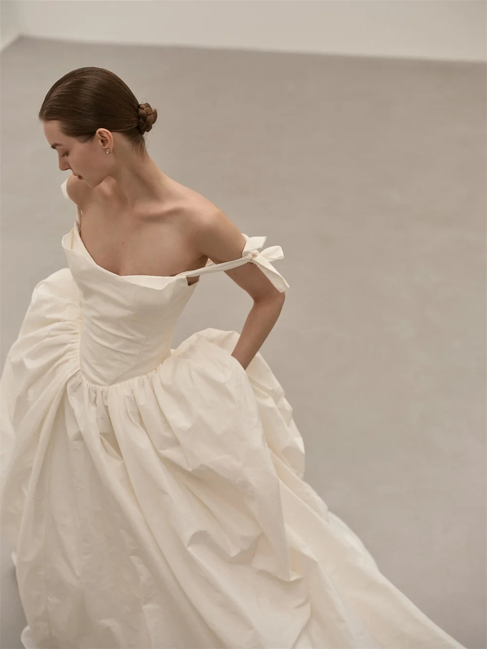 A woman wearing a voluminous off-the-shoulder white dress with a gathered bodice and flowing skirt, photographed in a minimalist light gray studio setting.