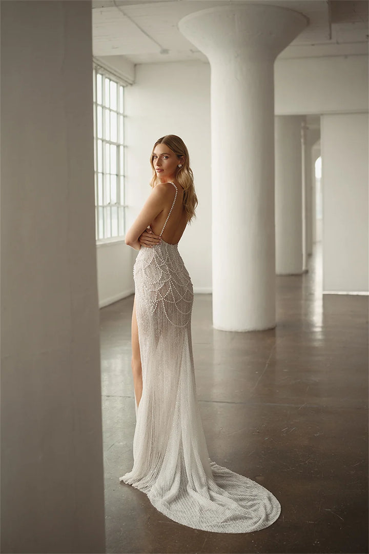A woman wearing a fitted white lace wedding gown with thin straps, a high slit, and a long train, standing in a bright industrial loft with white columns and large windows.