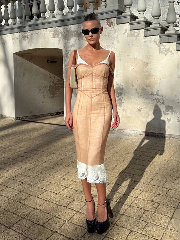 A woman stands outdoors wearing a fitted beige midi dress with white lace trim at the hem, white shoulder straps, black sunglasses, and black heels. The background shows a sunlit stone wall and tiled ground.