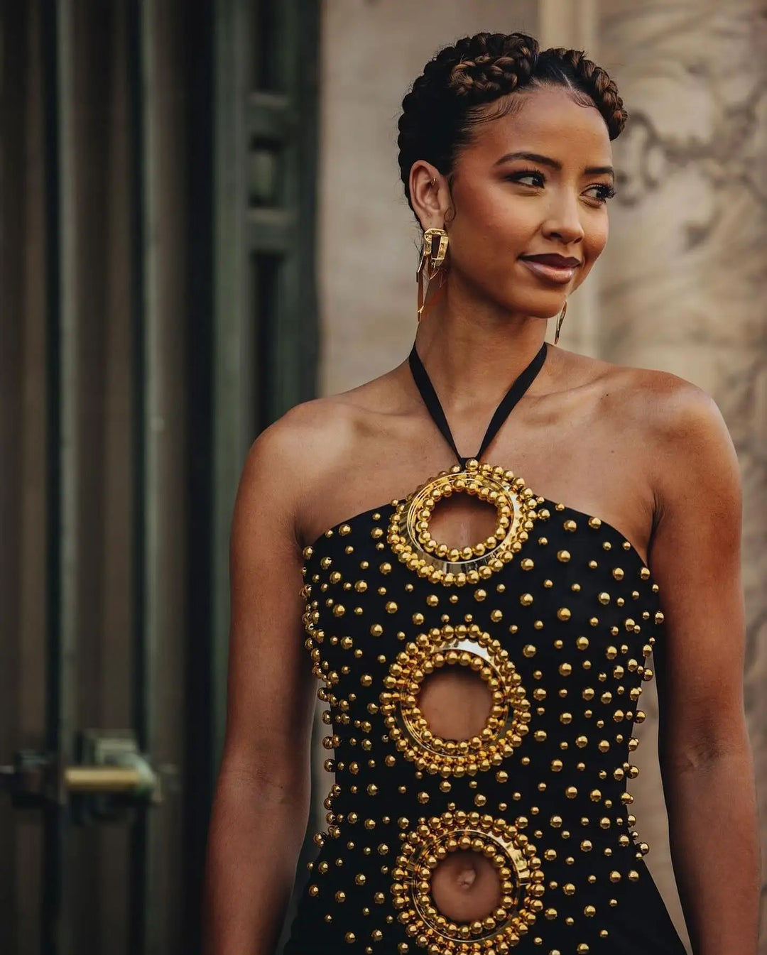 Woman wearing a black halter top with gold circular patterns against a stone wall.