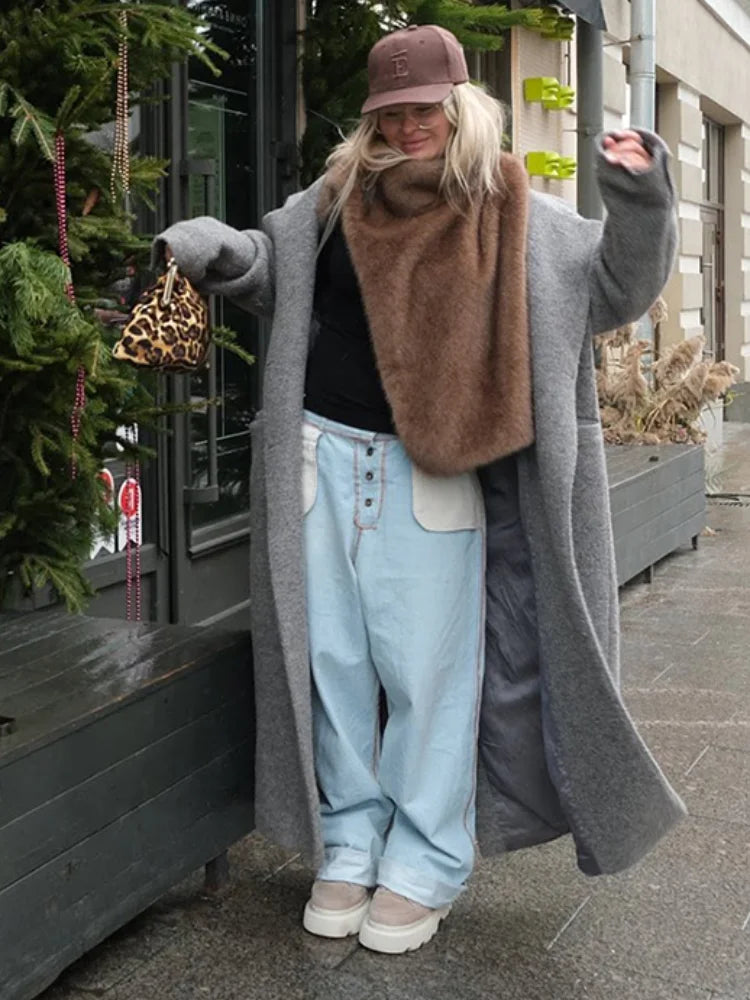 Person wearing a long gray coat, brown scarf, light blue jeans, and beige shoes on a city street.