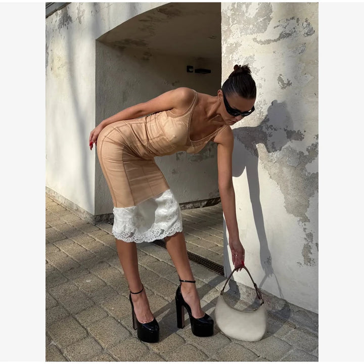 A woman wearing a sheer beige bodycon mini dress with a white lace hem, black platform high heels, and a white handbag, posing outdoors against a textured concrete wall.