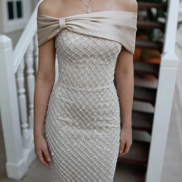 Beige off-shoulder dress with a large bow detail on a blurred staircase background