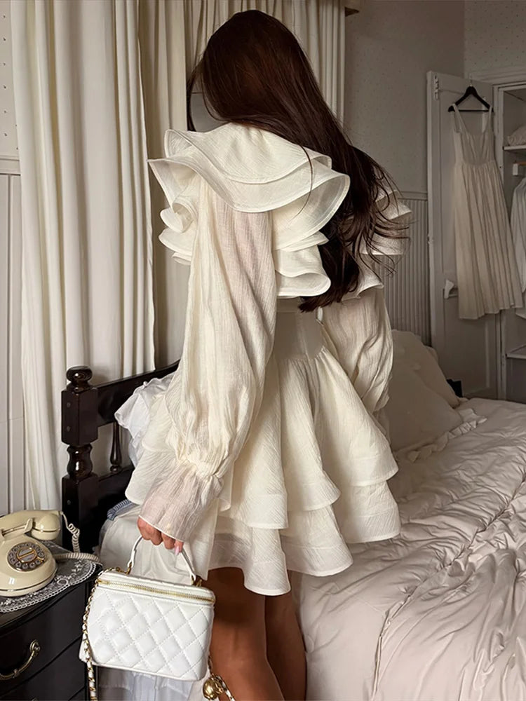 Woman in a white ruffled dress standing in a bedroom with a bed and dresser visible.