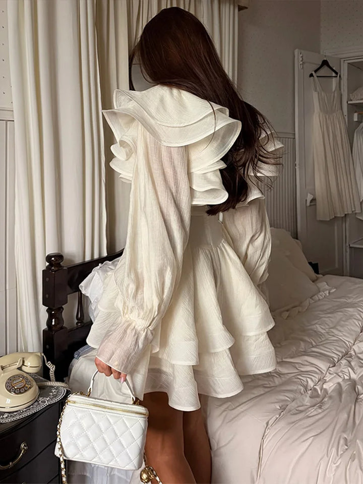 Woman in a white ruffled dress standing in a bedroom with a bed and dresser visible.