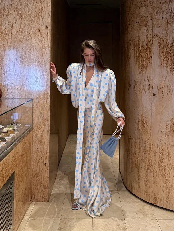 Woman in a long, patterned dress holding a blue handbag in a modern interior setting.