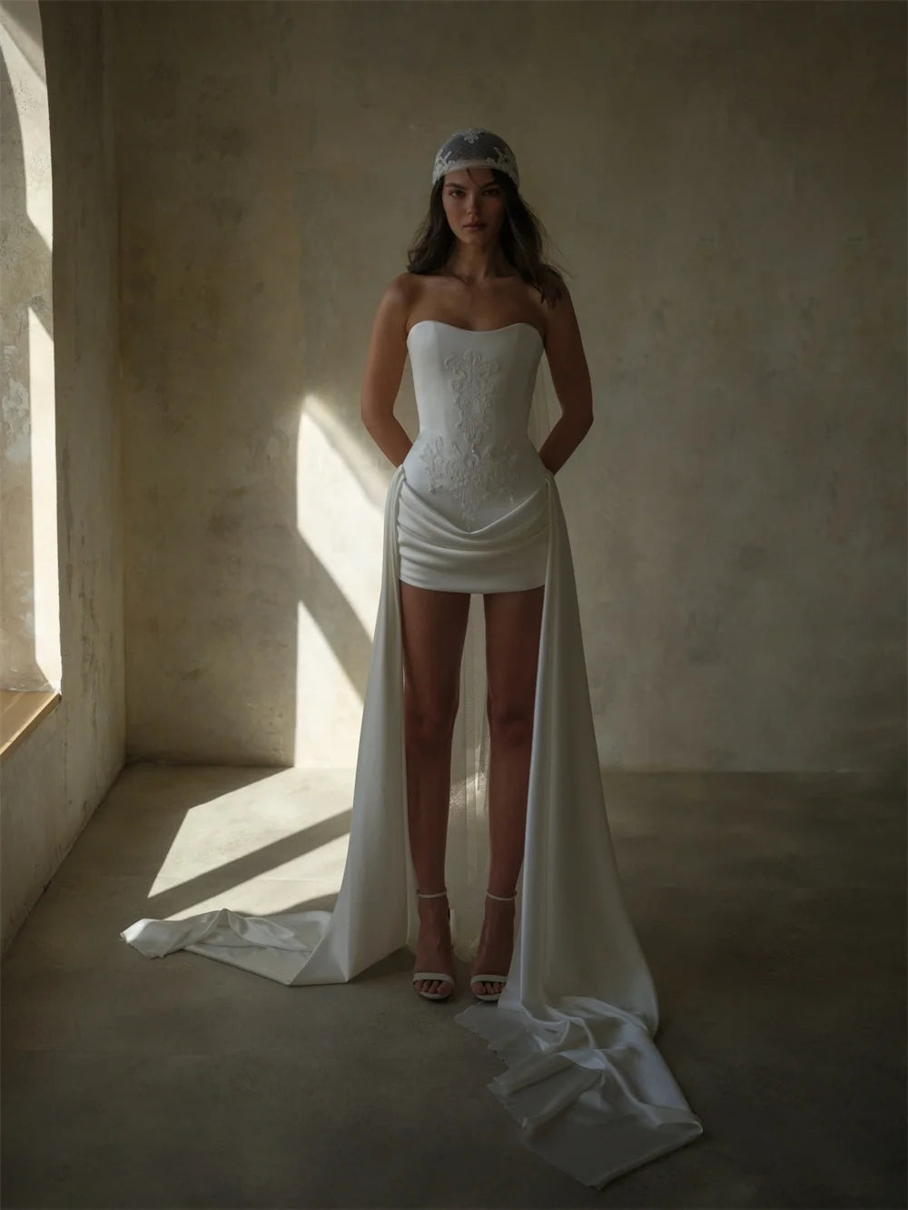 A woman stands in a sunlit room wearing a strapless white wedding dress with a fitted lace bodice, draped skirt, and long flowing train, paired with nude high-heel sandals and a headpiece.