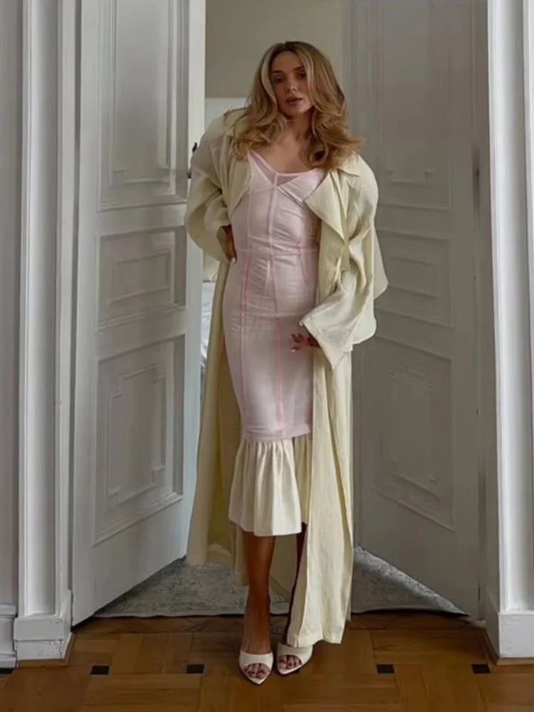 A woman stands in a doorway wearing a pale pink fitted midi dress with a ruffled hem, layered under a long cream-colored coat, paired with white heeled sandals.