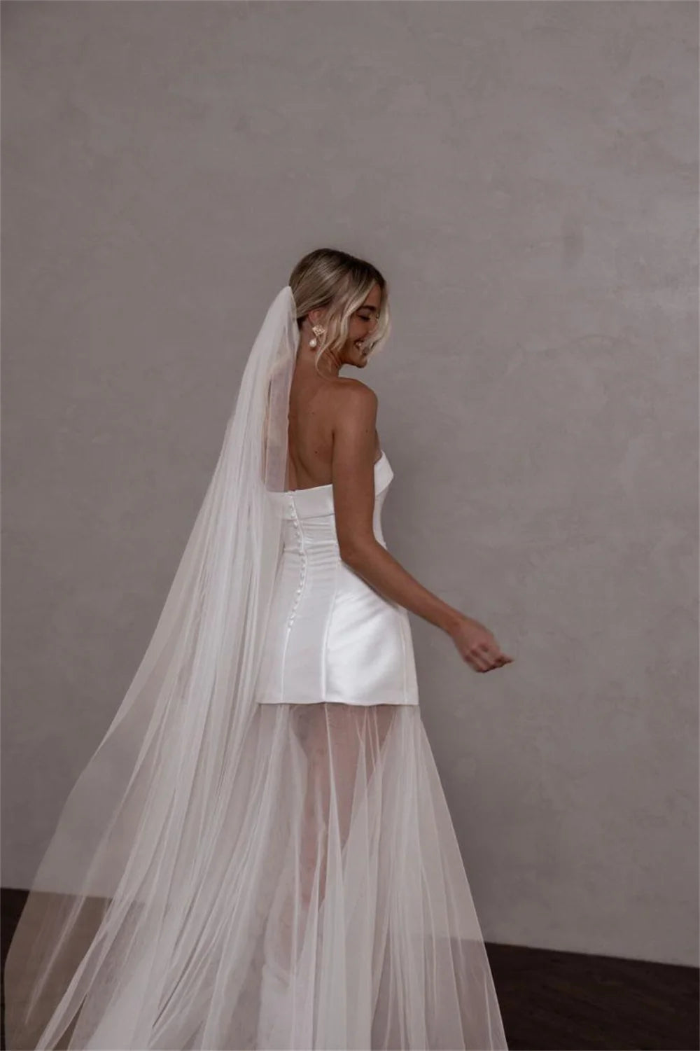 A woman wearing a white strapless wedding dress with a structured bodice, sheer tulle overskirt, and a long flowing veil, photographed against a plain textured gray wall.