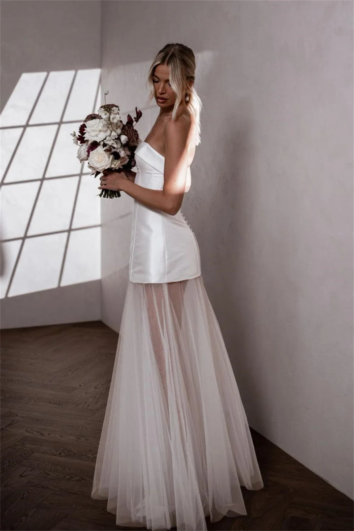 A woman wearing a strapless white wedding dress with a structured bodice and a flowing sheer tulle skirt, holding a bouquet in a minimalist room with sunlight streaming through a window.