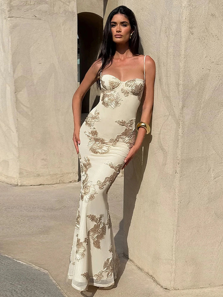 Woman in a white evening gown with gold floral patterns standing against a stone wall.