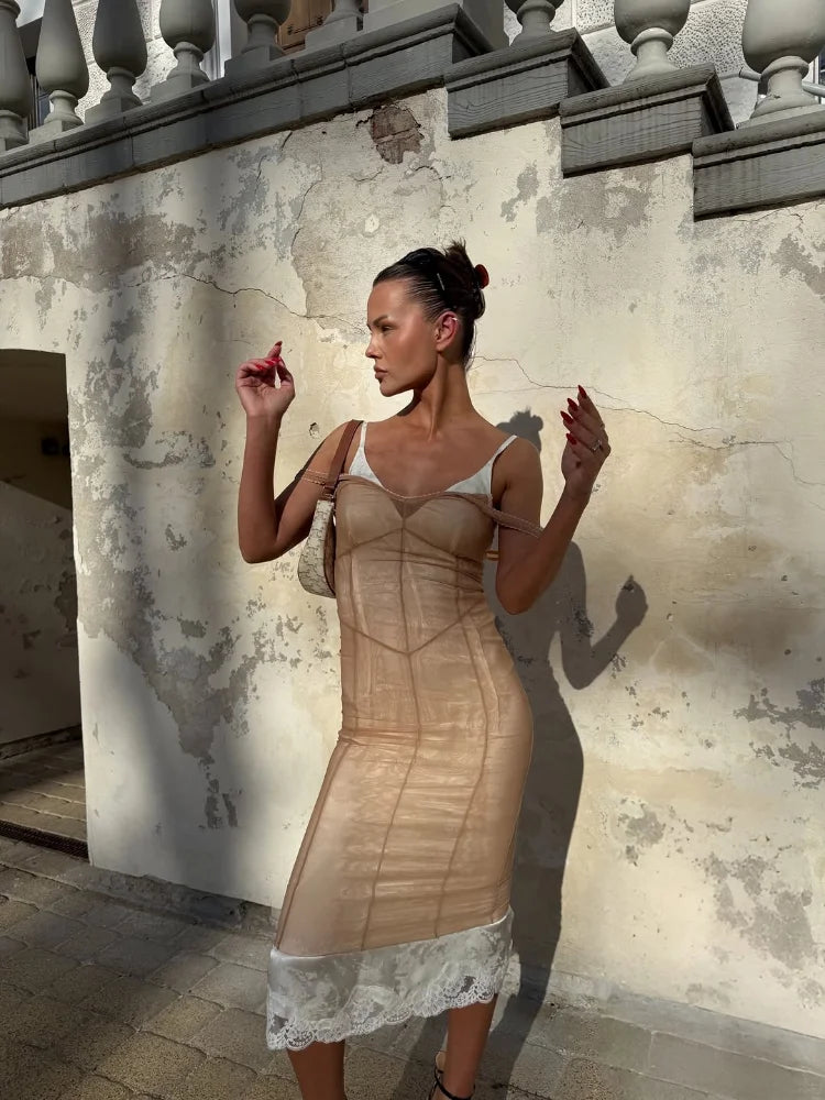 A woman stands outdoors against a weathered beige wall wearing a sheer nude-beige corset-style dress with white lace trim at the hem and white shoulder panels.