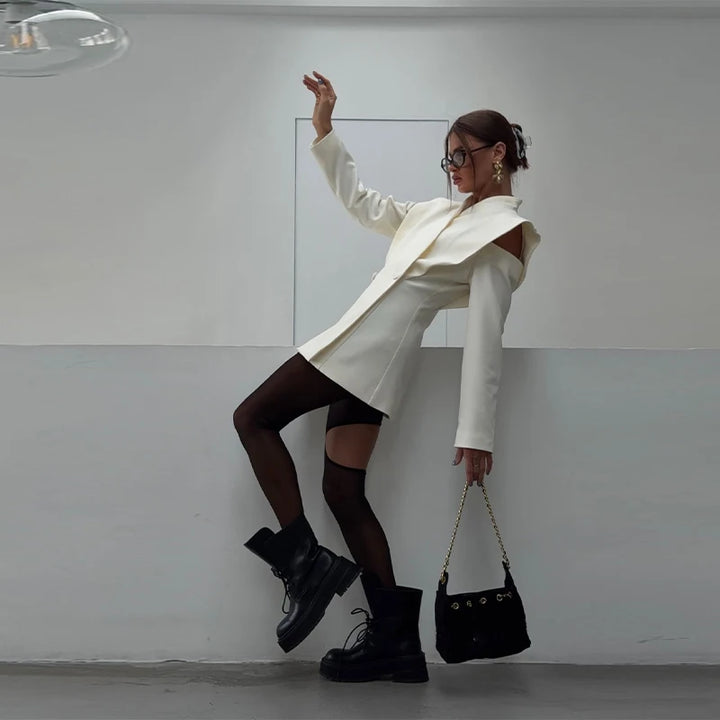Person wearing a white coat and black shoes, holding a black handbag against a plain background