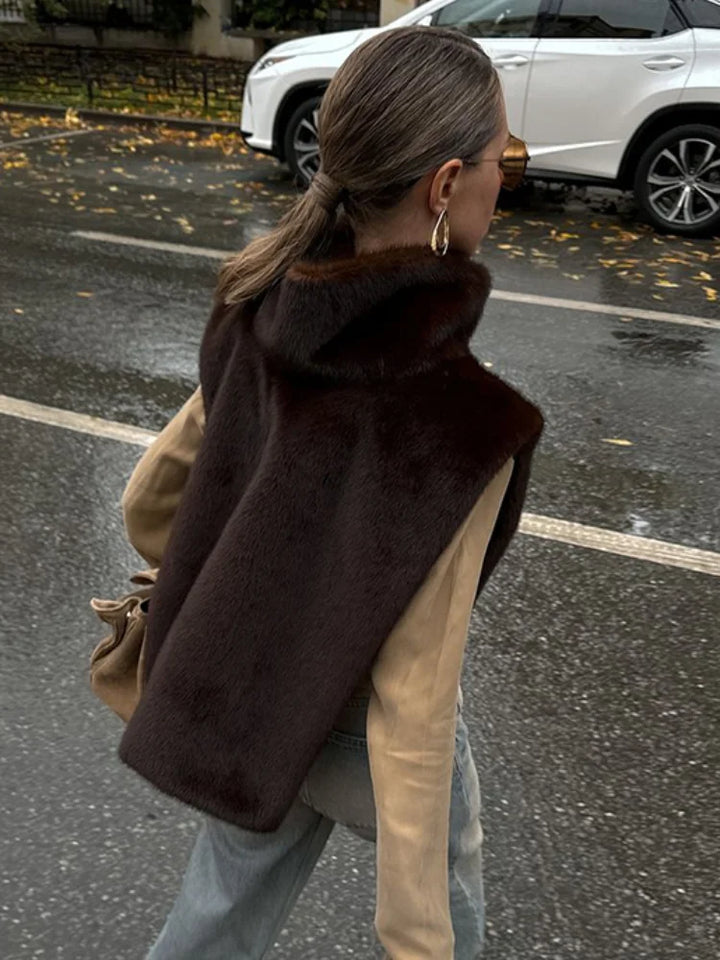 Person wearing a brown fur vest over a beige coat on a street.