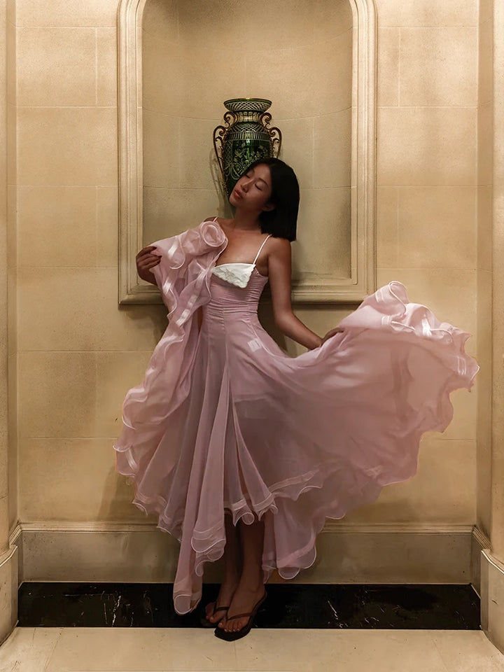 Woman in a pink dress standing in an elegant interior setting with a large mirror and decorative vase.
