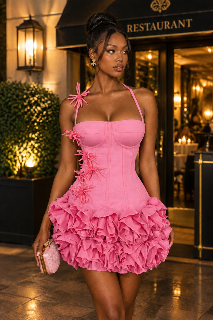 A woman models a bright pink mini dress with a structured corset-style bodice, halter neck, and a voluminous ruffled skirt, standing in front of a restaurant entrance at night.